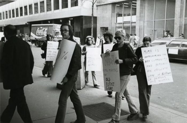 1976年，南希·斯佩罗在 MoMA 门口示威，她的牌子上写着：“MoMA 还在玩儿着精英主义的老把戏。”摄影：Mary Beth Edelson。图片致谢勒隆画廊