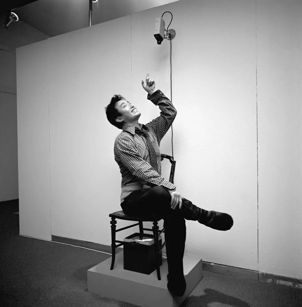 Nam June Paik sitting on TV Chair (1968/1976) in Nam June Paik Werke 1946–1976: Musik—Fluxus—Video, Kölnischer Kunstverein, Cologne, 1976. Photo: F. Rosenstiel, Cologne, Zentralarchiv des internationalen Kunsthandels (ZADIK), Cologne, Courtesy Gagosian.
